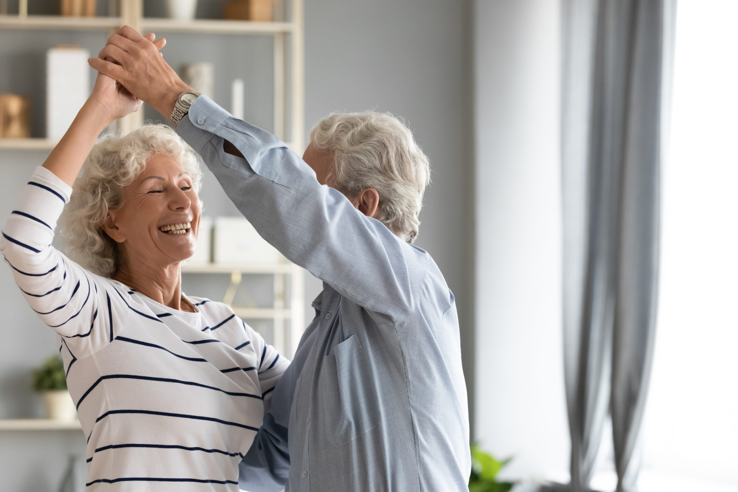Overjoyed elderly 60s couple spouse dance swirl in living room together, happy smiling mature retired husband and wife have fun engaged in funny activity, feel optimistic active enjoy weekend together